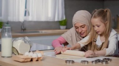 First bakery. Cute concentrated little girl pinning dough at kitchen, her loving muslim mother wearing hijab helping her, tracking shot, slow motion, empty space