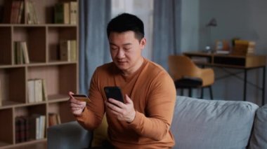 Çevrimiçi kazanma kavramı. Orta yaşlı Asyalı bir adam kredi kartı bilgilerini mobil uygulama üzerinden giriyor ve bankacılık işlemleri yapılıyor, evde başarılı bir işlemin keyfini çıkarıyor ve kutluyor, yavaş çekim