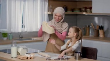 Happy muslim mother wearing traditional headscarf spinning dough on hand like professional pizzaiolo, teaching her little daughter to do the same, cooking together at kitchen, slow motion, free space