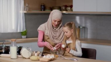 Family culinary. Happy muslim mother teaching her cute little daughter to cook at home kitchen, pretty girl cracking egg into bowl, slow motion, free pace