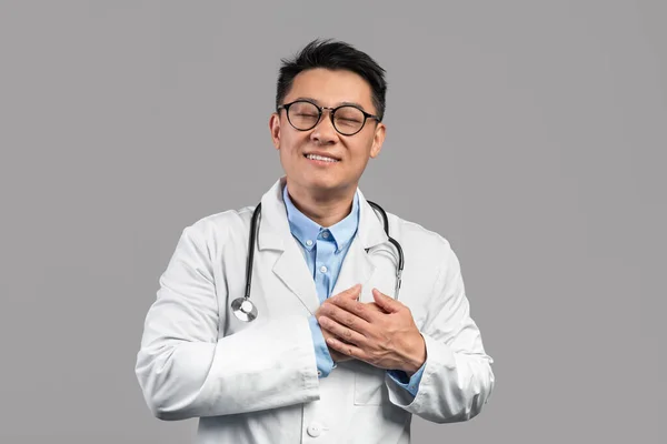 Happy millennial korean guy doctor in white coat, glasses with stethoscope presses hands to heart and thanks patient, isolated over gray studio background. Medicine and healthcare, grateful and hope