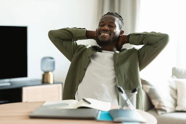 Happy african american man with closed eyes sitting at table and resting, leaning back at workplace, holding hands behind head, enjoying freelance job