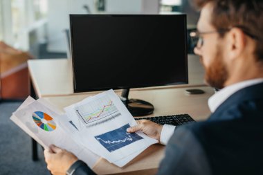 Successful entrepreneur reading financial reports, sitting at workplace with computer with black empty screen, space for mockup. Rear view, copy space