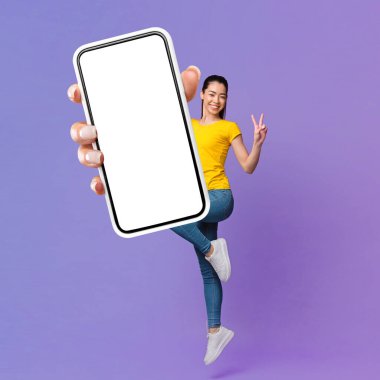Neşeli Güzel Asyalı Milenyum Kadını Havaya Atlıyor, Boş Ekran ve Barış Hareketi ile Akıllı Telefon Gösteriyor, Mor Arkaplan Üzerinde Tam Uzunluk, Boş Alan, Mockup, Kolaj