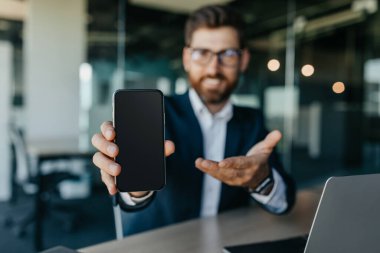 Happy businessman showing smartphone with blank screen, sitting at modern office, mockup for mobile app or website. Male entrepreneur in suit showing blank cellphone with copy space
