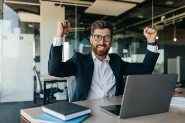 Emotional businessman gesturing yes and shaking fists, sitting at workplace in office and smiling at camera. Entrepreneur celebrating great deal. Successful career concept
