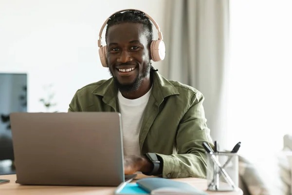 Positive black man in wireless headphones working online on laptop at home office, looking and smiling at camera, copy space. Happy guy communicating online on portable pc