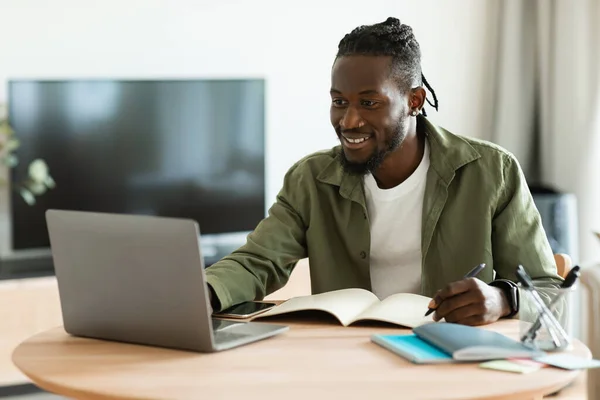 Excited black guy looking for job online, sitting at table, using laptop computer and taking notes, copy space. Positive african american man working online from home, remote job