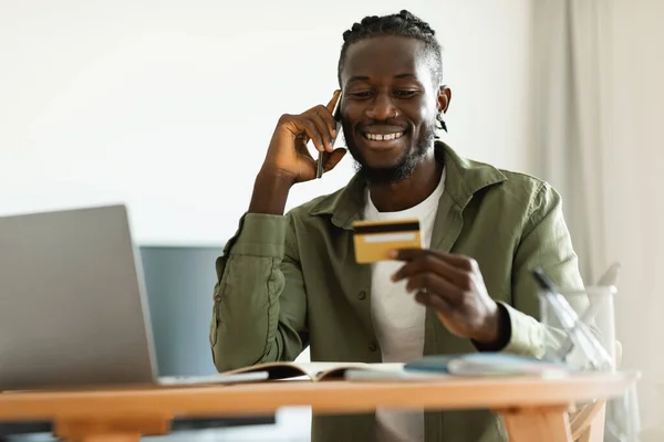 Excited black man holding credit card, having phone conversation with bank representative, sitting at home, copy space. Happy african american guy paying online