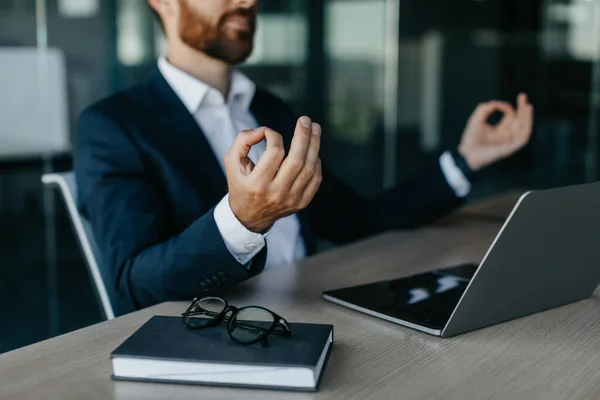 Relaxed businessman meditating at workplace in office, calm male entrepreneur in suit sitting at desk and coping with stress at work, selective focus