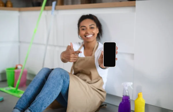 Happy millennial african american woman housewife sit on floor with cleaning supplies, pointing finger at smartphone with blank screen in kitchen interior. Home cleaning service, ad, app for homework
