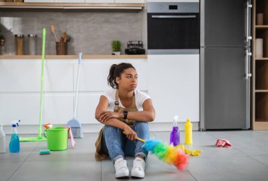Bored tired millennial black housewife sit on floor with cleaning supplies and brush, looking at empty space in kitchen interior. House cleaning service, many household chores, problems with housework