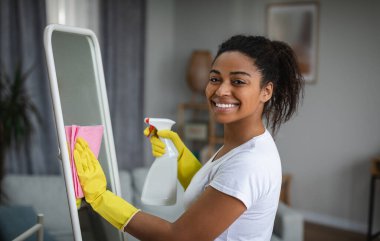 Satisfied millennial african american woman housewife in rubber gloves washing mirror with spray and microfiber rag in room interior. Household chores, house cleaning, housewife and perfect tidiness