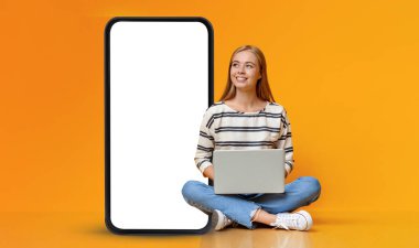 Dreamy teenage girl sitting with laptop on floor and looking at huge mobile phone with white empty screen, checking educational mobile app, orange studio background, mockup, collage, copy space