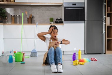 Shocked young black housewife with open mouth sit on floor with cleaning supplies, look at clock in kitchen interior. Late with cleaning, house cleaning service, a lot of household chores and chaos