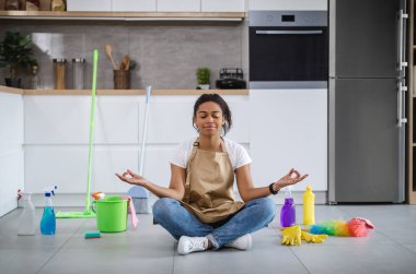 Smiling calm millennial african american woman housewife with closed eyes sitting on floor with cleaning supplies, meditating in kitchen interior. Mental health care, house cleaning, household chores
