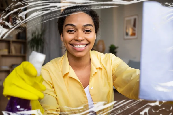 Happy Black Woman Cleaning Window Glass With A Rag And Detergent Smiling To Camera Wearing Yellow Rubber Gloves At Home. Woman Doing Housework. Selective Focus
