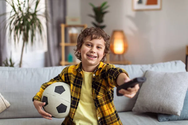 Sport supporter. Little boy football fan watching tv at home, sitting on sofa with remote controller and soccer ball, smiling to camera