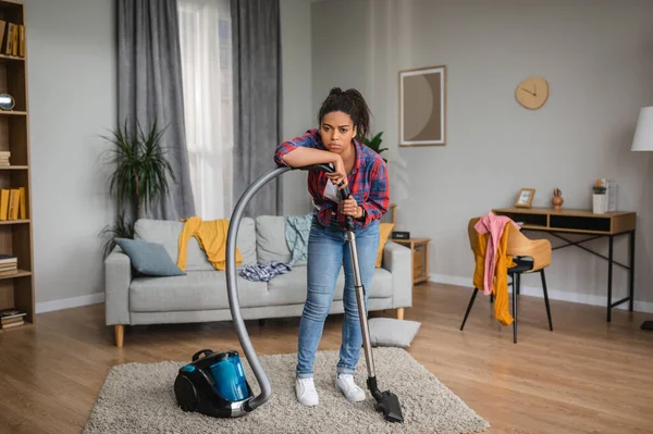 Unhappy tired sad millennial african american housewife vacuuming and rest from cleaning in living room interior. Household chores, troubles in domestic work, disorder at home, chaos with clothes