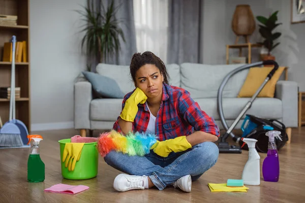 Boring thoughtful millennial african american housewife in rubber gloves with cleaning supplies sit on floor with brush in cozy living room interior. Cleaning, household chores and disorder at home