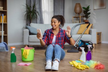 Cheerful millennial african american housewife waving hand at smartphone sitting on floor in living room interior with cleaning supplies. Video call during housework, household chores and gadget