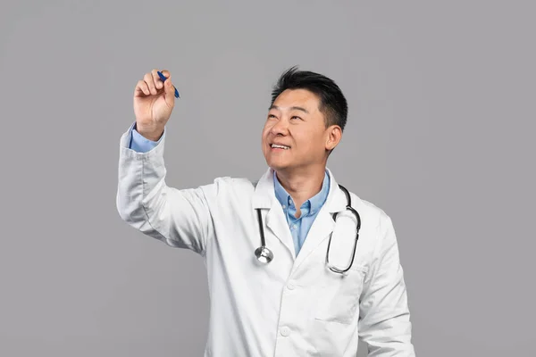 Smiling adult asian guy doctor in white coat with stethoscope writes on virtual blackboard with blank space isolated on gray background. Modern technology for medicine, health care and diagnostics