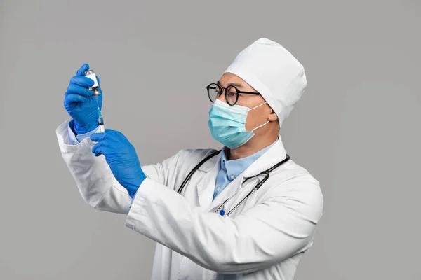 Busy serious adult korean doctor in white coat, glasses and gloves take vaccine into syringe, isolated on gray background, studio. Vaccination and immunization from flu and covid-19 virus and medicine
