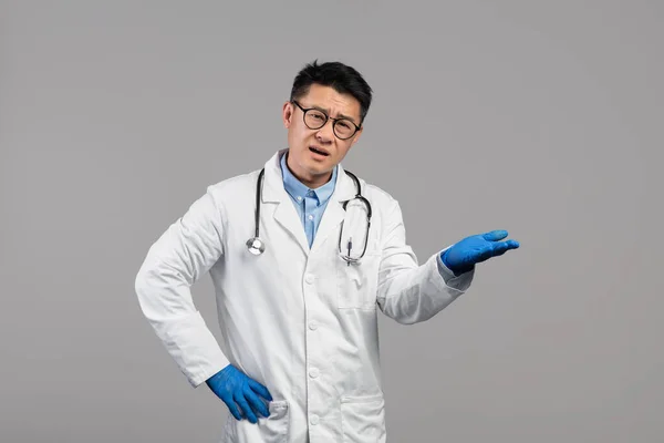 Sad confused adult asian man doctor in white coat, goggles and protective gloves holds empty space on his hand, gesticulate isolated on gray background, studio. Health care and problems in medicine