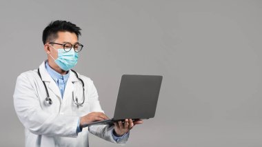 Confident adult chinese man doctor in white coat, glasses, protective mask typing on laptop isolated on gray background. Medicine, health care, social distance and online consultation due covid-19