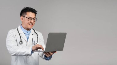 Beyaz önlüklü, güveni tam orta yaşlı Asyalı doktor, steteskoplu gözlükler gri arka planda, stüdyoda dizüstü bilgisayarda daktilo ediyor. Online danışma, tıbbi servis kumandası, sağlık hizmetleri