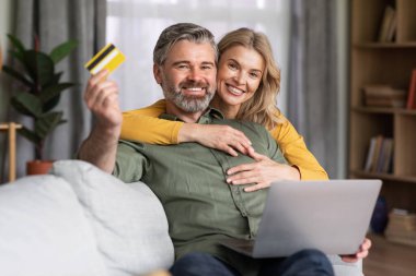 Online Payments. Happy Middle Aged Spouses Posing With Laptop And Credit Card In Home Interior, Smiling Married Couple Sitting On Couch In Living Room, Recommending Internet Purchases, Copy Space