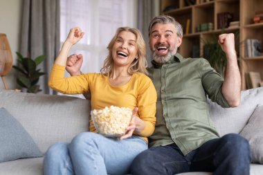 Domestic Leisure. Excited Middle Aged Spouses Watching Sports On Tv At Home, Married Couple Eating Popcorn And Cheering Favorite Team While Relaxing On Couch In Living Room, Raising Fists With Joy