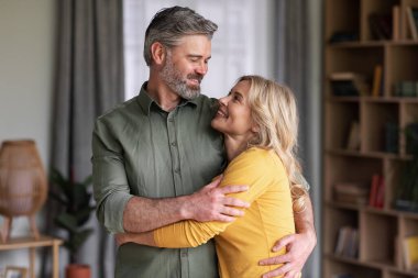 Evde kucaklaşan ve gülümseyen romantik orta yaşlı bir çiftin portresi, mutlu eşlerin oturma odasında dinlenirken kucaklaşmaları, boş zamanların tadını çıkarmaları, boşluğu kopyalamaları.