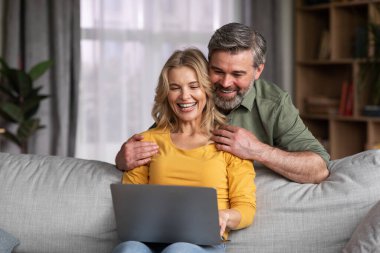 Mutlu orta yaşlı çift dizüstü bilgisayarlarıyla evde eğleniyor, keyifli eşler oturma odasında dinlenirken bilgisayar kullanıyor, internete bakıyor, internetten alışveriş yapıyor, mekanı kopyalıyor.