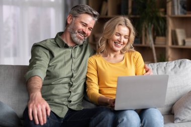 Çevrimiçi Hobi. Evde dizüstü bilgisayar kullanan gülümseyen orta yaşlı eşler, mutlu koca ve eş oturma odasında bilgisayarla dinleniyor, sosyal medyaya bakıyor ya da internetten alışveriş yapıyor,