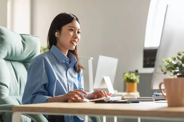 Happy Asian Millennial Businesswoman Working Online On Computer Typing On Keyboard Browsing Internet Wearing Earbuds Earphones Sitting In Modern Office, Side View. Business Career Concept