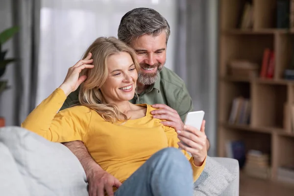 Happy middle aged couple using smartphone while relaxing on couch at home, positive spouses watching photos or browsing internet on mobile phone while resting together in living room, closeup shot