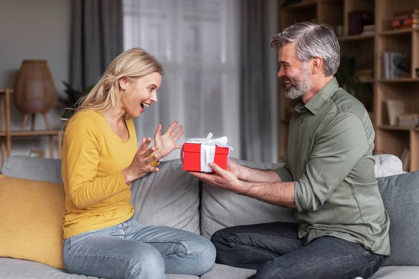 Loving middle aged husband surprising his wife with gift at home, greeting her with birthday or anniversary, happy man giving present box to excited spouse, celebrating valentines day together