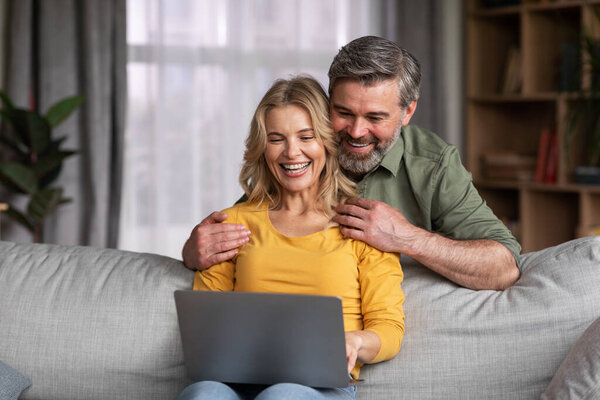 Happy Middle Aged Couple With Laptop Having Fun Together At Home, Cheerful Spouses Using Computer While Relaxing On Couch In Living Room, Browsing Internet Or Shopping Online, Copy Space