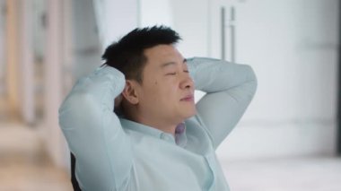 Rest at work. Close up portrait of pensive middle aged asian businessman relaxing at workplace, swinging on chair at office, slow motion