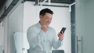 Great news concept. Indoors portrait of happy middle aged asian businessman reading email on smartphone and celebrating win, standing in office, slow motion