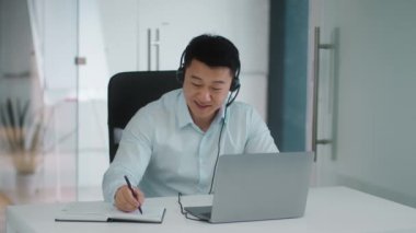 Corporate virtual meeting. Happy middle aged asian man employee in headset video chatting with colleagues online via laptop, taking notes in office and laughing, slow motion