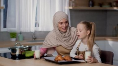Healthy homemade lunch. Happy little girl and her muslim mother in hijab smelling fresh hot croissants and enjoying milk in glasses at kitchen, slow motion, empty space