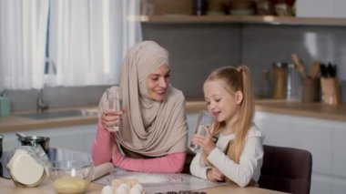 Inculcation of good habits. Portrait of happy muslim mother wearing hijab and her little daughter drinking water from glass at kitchen, laughing together, slow motion