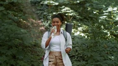 Active weekends. Young sporty african american woman tourist enjoying walk in forest, drinking water from bottle, hiking with backpack in woodland, follow shot, slow motion