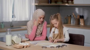 Preparing homemade cookies. Curious little girl cutting dough with metal cutters at kitchen, loving muslim mother wearing traditional headscarf watching her daughter, slow motion, free space