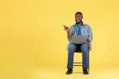 Neşeli Afro-Amerikalı Adam Laptop Bilgisayarlı Parmak Yanı Reklam Sitesi ya da Sarı Stüdyo Arkaplanı Üzerinde Oturan Online Teklif Kullanıyor. İnternet ve Teknoloji. Boşluğu Kopyala