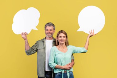Happy middle aged spouses holding empty speech bubbles on yellow studio background, mockup for design. Positive european man and woman with blank word clouds