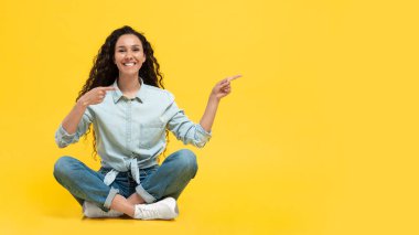 Happy Middle Eastern Woman Pointing Fingers Aside Showing Free Space Smiling To Camera Sitting In Studio Over Yellow Background. Female Advertising Yout Text. Look There. Panorama