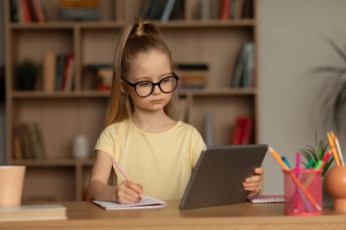 E-öğrenme. Ciddi liseli kız dijital tablet kullanıyor ödevlerini yapıyor ve evde not alıyor. Çevrimiçi Öğrenen Çocuk Bilgisayarda Video Dersi İzliyor, Gözlük takıyor